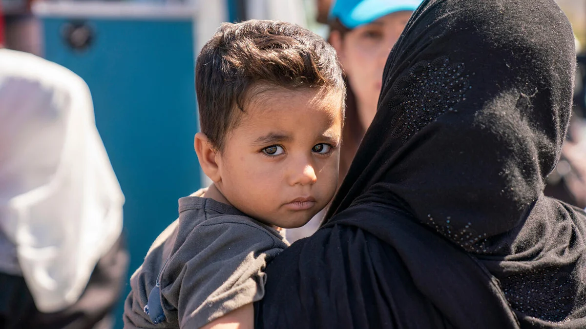 Kind schaut in die Kamera und wird von Frau im Arm gehalten. Im Hintergrund sieht man eine UNICEF-Mitarbeiterin.