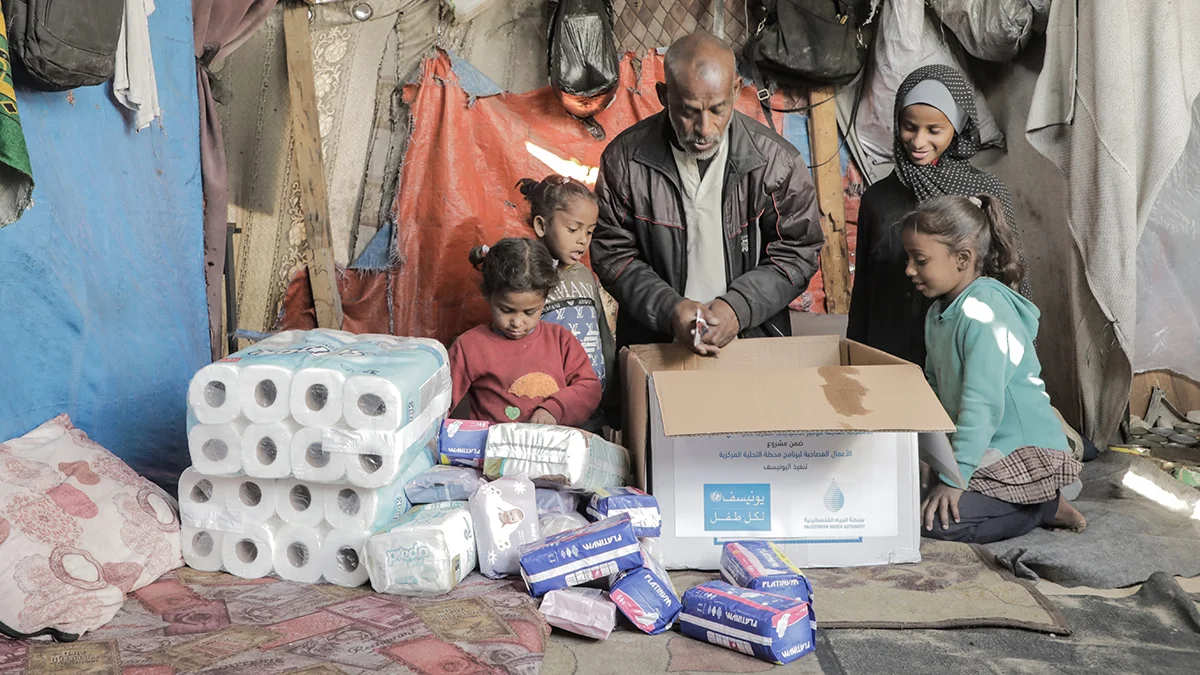 Ein Vater sitzt mit seinen Kindern in ihrer Notunterkunft vor einem UNICEF Karton, der mit Hygieneprodukten gefüllt ist.