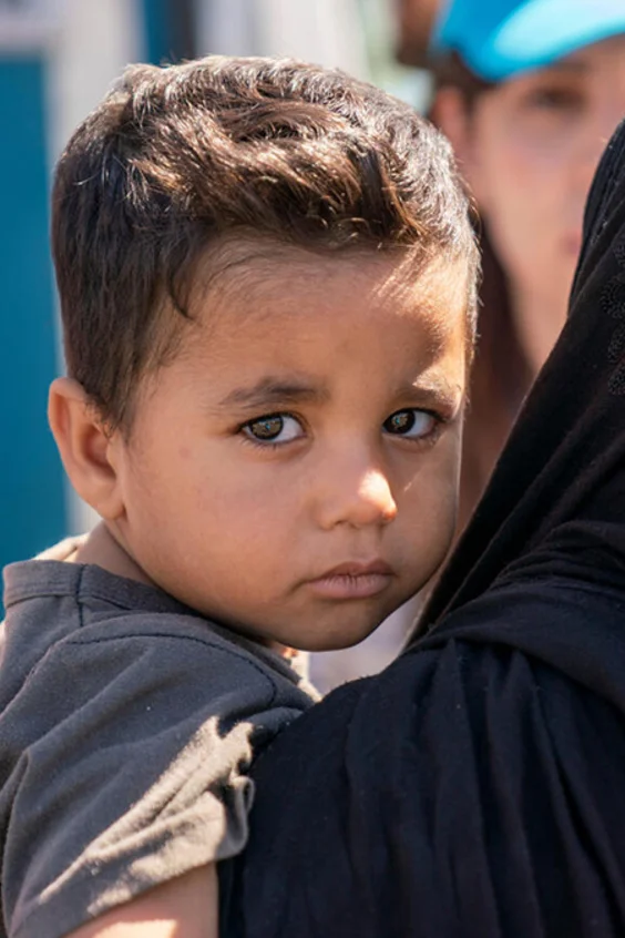 Kind schaut in die Kamera und wird von Frau im Arm gehalten. Im Hintergrund sieht man eine UNICEF-Mitarbeiterin.